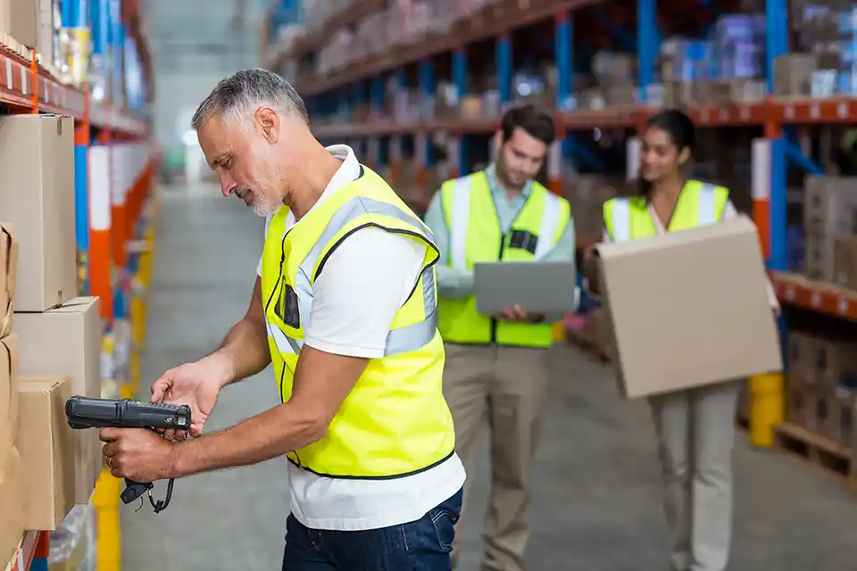 Tre personer med gula västar på ett lager. En man scannar med rfid scanner två andra logistik arbetare hämtar ut kartong för sändning.